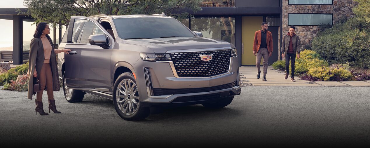 Cadillac Escalade sits in the driveway of a modern home. A woman stands at the open passenger side door while a man and a young boy walk out of the home toward the car. OnStar Insurance logo is in the bottom right corner.